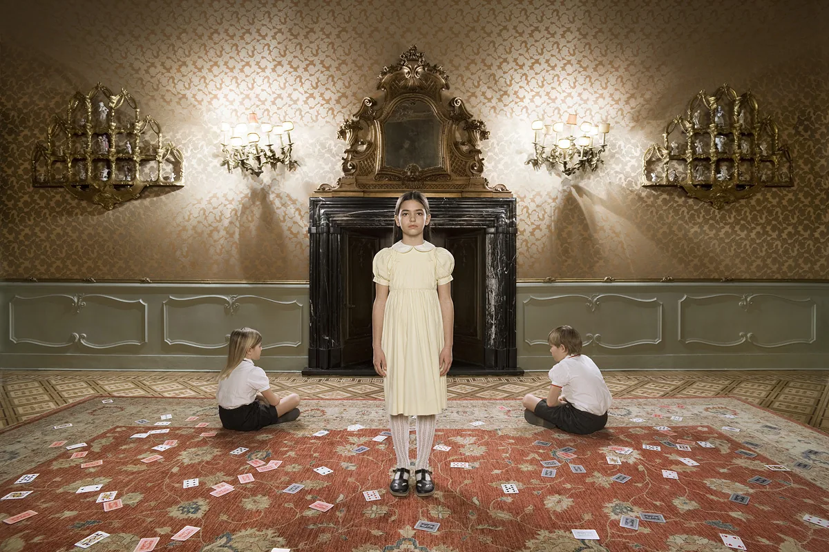 Bambina in abito giallo tra due bambini seduti, con carte da gioco sparse sul tappeto di una sala dorata — Filippo Piantanida e Roberto Prosdocimo