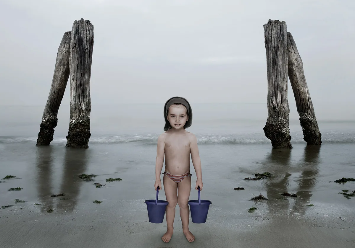 Bambino con cuffia e due secchielli su una spiaggia grigia tra due vecchi pali di legno, mare in foschia — Filippo Piantanida e Roberto Prosdocimo
