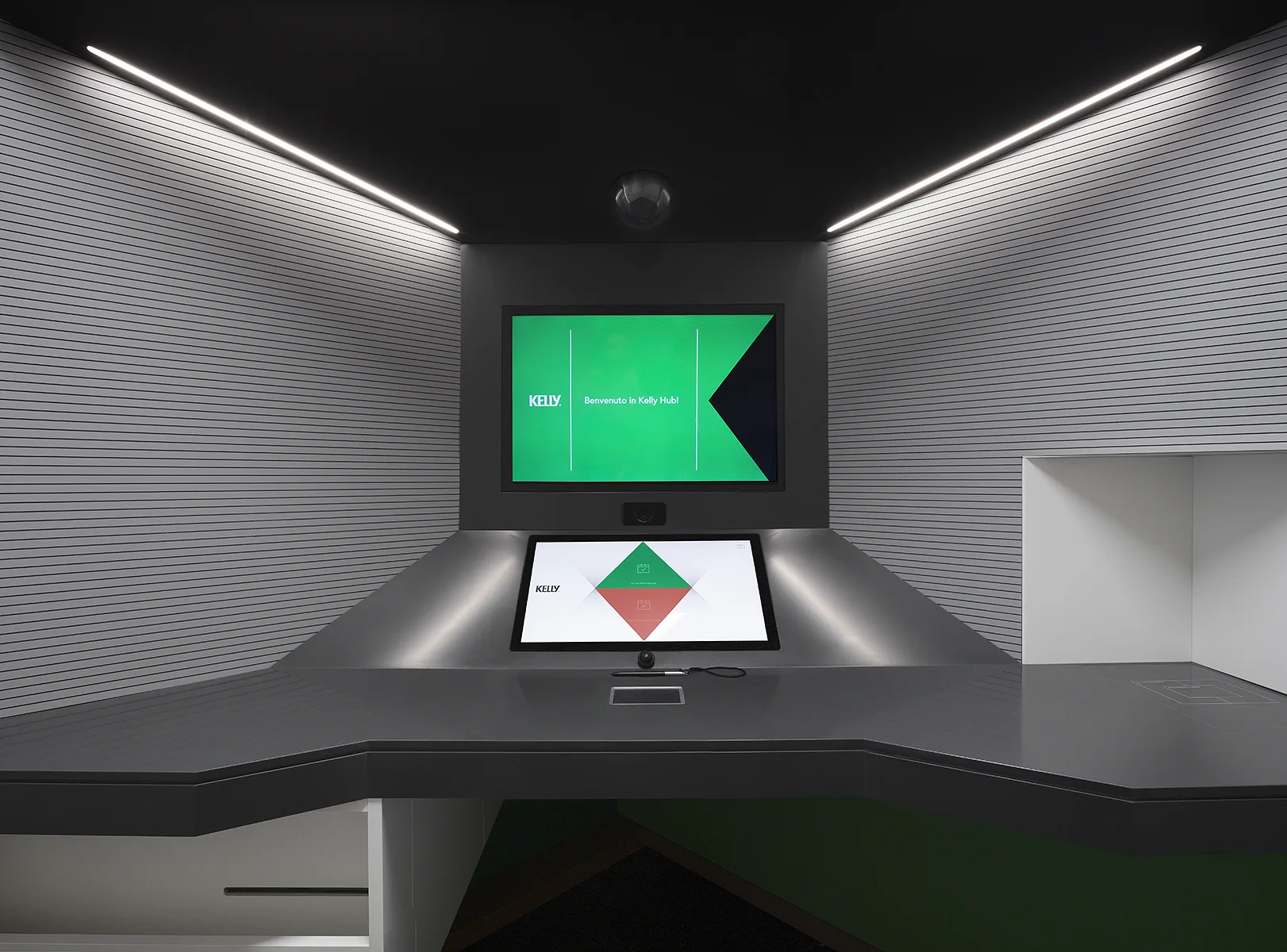 Vista frontale della postazione Kelly Hub con schermo di benvenuto verde e monitor touch sul piano grigio antracite, pareti a doghe — foto Filippo Piantanida