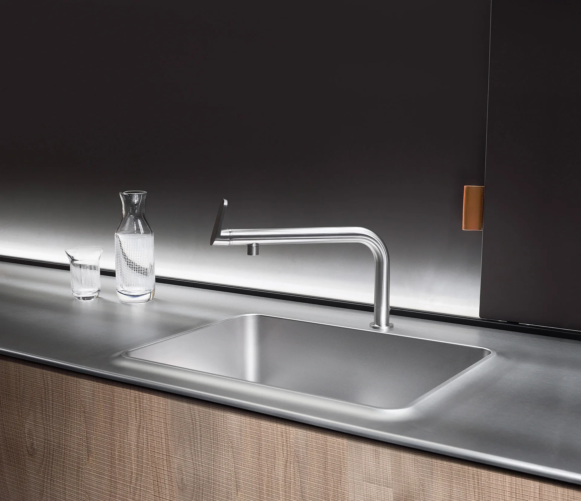 Steel sink with a brushed gooseneck tap, an engraved crystal carafe and glass, light wood door — Wallpaper editorial, Filippo Piantanida Studio